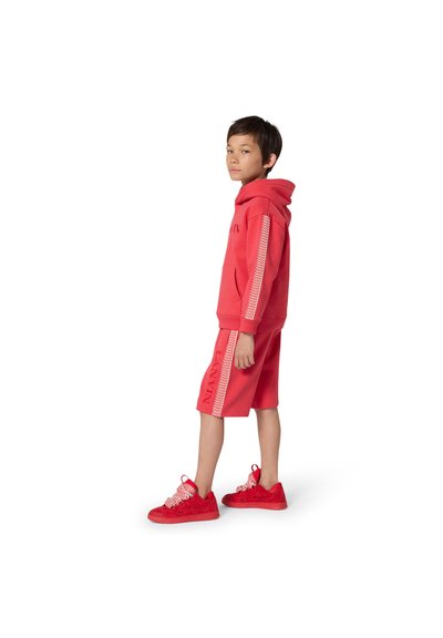 Jeune garçon debout, portant un sweat à capuche rouge et un short assorti avec des rayures à motifs, assortis à des baskets rouges.