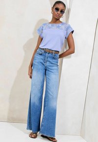 Tricou albastru deschis cu detalii din dantelă, asociat cu blugi din denim albastri cu picioare largi. Accesoriile includ ochelari rotunzi de soare și un curea.