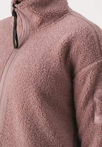 Chaqueta de forro polar rosa difusa con cuello alto y cierre con cremallera, que presenta un tejido texturizado y un parche en la manga.