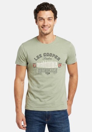 Homme souriant portant un t-shirt Lee Cooper London vert clair avec texte imprimé et un jean bleu, debout devant un fond uni.
