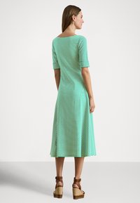 Nő zöld-fehér csíkos midi ruhában, könyékig érő ujjakkal, barna ék sarkú szandált viselve, egy egyszerű háttér előtt oldalra fordulva áll.