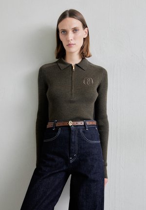 Pulôver verde-oliva canelado com zíper de um quarto e colarinho; apresenta um logótipo no peito. Combinado com jeans escuros de cintura alta e um cinto castanho.