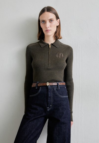 Ribbet oliven grøn pullover med kvart lynlås og krave; har et logo på brystet. Kombineret med højtaljede mørke denim jeans og et brunt bælte.