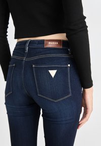 Tmavomodré džíny z denimu s priliehavým strihom, vybavené hnedou koženou etiketou s nápisom "GUESS", zlatým prešívaním a trojuholníkovým logom na zadnej vrecku.