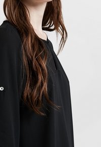 Musta pusero, jossa on väljää istuvuutta, pieni avainreikäkaulus ja napitus hihassa. Sileä pinta.