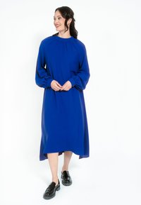 Langärmliges blaues Kleid mit hohem Kragen, Raffung an den Schultern und lockerer Passform, die bis zum knieumspielenden Saum verläuft. Schwarze Slipper getragen.