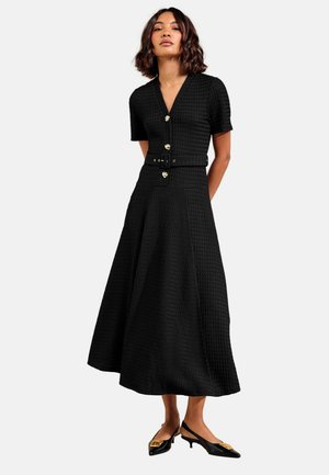 Kvinde iført en sort tekstureret midikjole med korte ærmer, guldknapper, bælte i taljen og sorte slingback kitten heel sko med gulddetalje.
