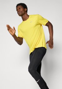 T-shirt Nike jaune à manches courtes, associé à des leggings noirs moulants avec un logo Nike blanc. Texture de tissu lisse.