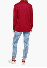 Person med en rød langærmet skjorte, blå jeans med palmebladers mønster, opsmøgede ærmer og hvide sneakers med røde metalhæle, der vender væk.
