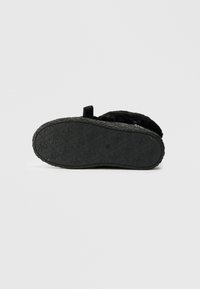 Black slipper with a soft, textured interior and an anti-slip rubber sole featuring a patterned tread design. Adjustable strap on the front.