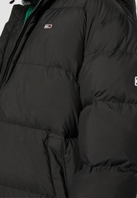 Svart puffjacka tillverkad av slät tyg, med dragkedja, sidofickor och en liten logotyp på bröstet.