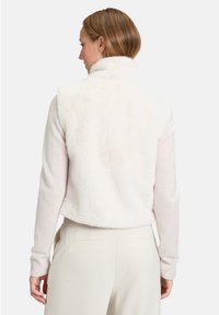Kamizelka z sztucznego futra w kolorze kremowym z wysokim kołnierzem, w połączeniu z lekkim swetrem. Miękka tekstura i krótki krój dobrze pasują do dopasowanych spodni.