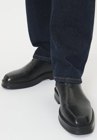 Čierne kožené členkové topánky so štvorcovou špičkou, s hladkým povrchom a robustnou podrážkou, skombinované s tmavomodrými džínsy.