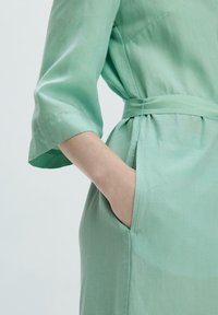 Mintgrünes Kleid mit Gürtel, das über drei Viertel-Ärmel, eine seitliche Tasche und eine glatte, leichte Stofftextur verfügt.