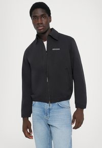 Black bomber jacket with a zip front and collar, featuring elasticized hem and cuffs. Small logo on the chest. Paired with light blue jeans.