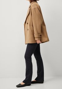 Bruine oversized petticoat met grote knopen, zijzakken en opgerolde mouwen; gecombineerd met uitlopende donkere jeans en zwarte flats.