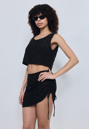 Femeie purtând un top negru fără mâneci, texturat, scurt și o fustă mini cu șnururi adunate, pozând cu o mână pe șold, purtând ochelari de soare negri.