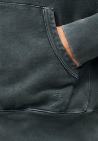 Χέρι ατόμου μέσα στην μπροστινή τσέπη σκούρας γκρι φούτερ με ριμπ μανσέτα και διακριτικό κεντημένο λογότυπο «MN».