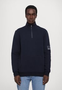 Sudadera de color azul marino con cuello alto, media cremallera, bolsillo frontal y logo bordado en la manga. Textura suave.