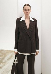 Blazer cruzado de rayas marrón con un gran cuello blanco, llevado sobre una camisa blanca. La modelo sostiene un bolso marrón oscuro, de pie sobre una superficie clara.