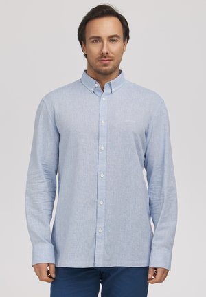 Mężczyzna stoi zwrócony przodem, ubrany w jasnoniebieską koszulę na guziki z długimi rękawami i ciemnoniebieskie spodnie.