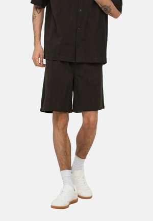 Adulto vestido con camisa negra abotonada y pantalones cortos a juego, calcetines blancos y zapatillas blancas sobre un fondo liso.