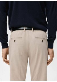 Pantalones beige de corte entallado, con parte delantera plana y dos bolsillos traseros con botones negros. Combinados con un suéter azul marino y un cinturón negro.