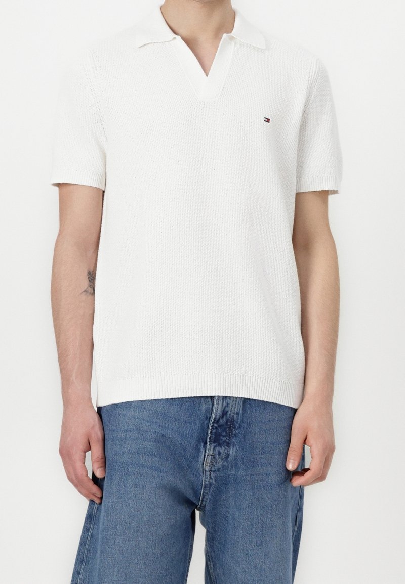 Homme portant un polo en maille blanc à manches courtes avec un petit logo sur la poitrine et un jean en denim bleu sur fond blanc.
