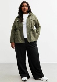 Leichtgewichtige olivgrüne Jacke mit Kapuze, zwei Klapptaschen, elastischem Bund und Reißverschluss, kombiniert mit einem weißen Shirt und schwarzen Hosen.