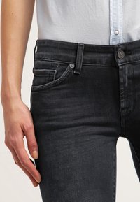 Los jeans de mezclilla gris oscuro presentan un corte ajustado, costuras claras y bolsillos delanteros con cierre de botón. La cinturilla incluye pasadores para el cinturón.