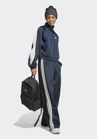 Chándal azul marino con acentos blancos, que incluye una chaqueta con cremallera y pantalones de pierna ancha. Combinado con una mochila negra y zapatillas blancas.