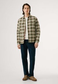 Camisa de franela a cuadros verdes con dos bolsillos en el pecho, camiseta blanca debajo, pantalones azul oscuro y zapatillas marrones con franjas laterales.