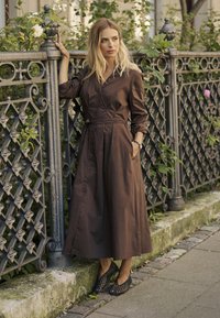 Sötétbarna, könnyű pamut ruha hosszú ujjakkal és �övvel a deréknál, fekete, szőtt cipőkkel párosítva; egy díszes fékfence mellett áll.