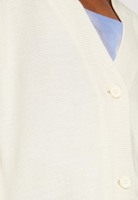 Nära bild av en vit ribbad cardigan med knappar över en blå skjorta på en person med brun hy.