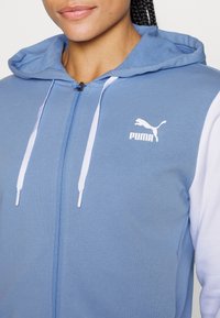 Femme portant un sweat à capuche bleu zippé avec manches blanches et un logo blanc Puma sur la poitrine, montrant le haut du torse et le cou.