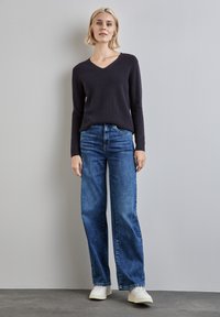 Navyblauer V-Ausschnitt-Pullover aus weichem Stoff, kombiniert mit weit geschnittenen blauen Jeans und weißen Sneakers vor einem grauen Hintergrund. Sauberer, lässiger Look.