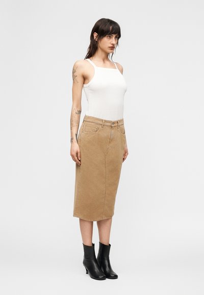 Γυναίκα με τατουάζ, ντυμένη με λευκό αμάνικο τοπ, καφέ μεσαίο φούστα και μαύρα μποτάκια στον αστράγαλο, στέκεται μπροστά σε μια απλή γκρι φόντο.