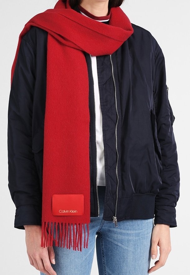 Personne portant une écharpe rouge vif avec des franges et une étiquette Calvin Klein sur une veste noire et un jean bleu clair, debout devant un fond blanc.
