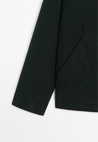 Dark green, long-sleeve garment with a smooth texture, featuring a front pocket and clean stitching. The sleeve has a straight cut.