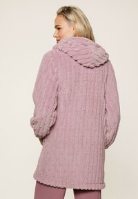 Abrigo texturizado rosa con capucha, que presenta un patrón de rayas verticales y suaves detalles de pompones. Mangas largas y diseño de longitud media.