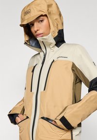 Wasserfeste Jacke in Tan und Creme mit schwarzen Akzenten, ausgestattet mit Reißverschlusstaschen, verstellbarem Kapuzen und strukturiertem Material für eine verbesserte Haltbarkeit.