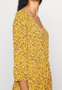Robe florale jaune avec un décolleté en V profond, de longues manches ballon, une taille froncée, et un tissu texturé orné de petites fleurs blanches et rouges.