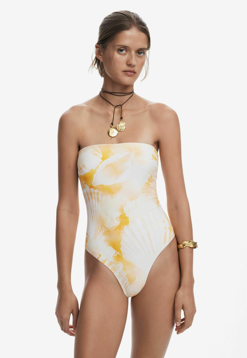 Maillot de bain sans bretelles arborant un imprimé coquillage blanc et jaune, avec une texture lisse et une coupe haute au niveau des jambes. Accessoirisé par un collier de choker doré.