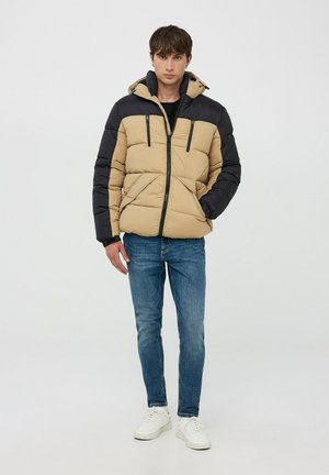 Giacca a vento beige e nera con tasche con zip, dotata di cappuccio, texture trapuntata e design aderente. Abbinata a jeans blu e sneakers bianche.