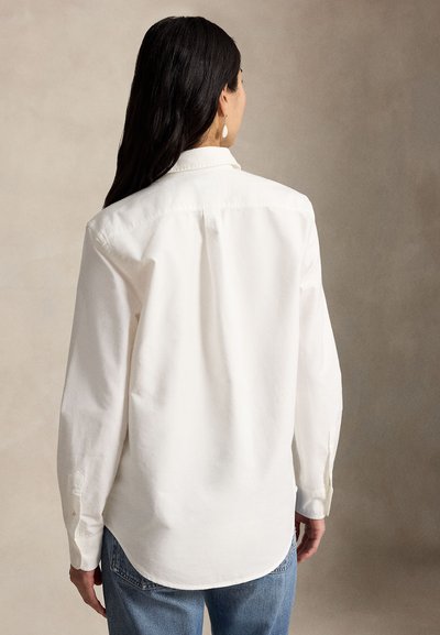 Femme aux longs cheveux foncés portant une chemise blanche à manches longues et un jean bleu, tournée de dos sur un fond neutre.
