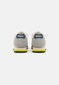 Sneakers beige e grigi in materiale scamosciato, con un logo blu sul tallone e un accento giallo sulla suola. Vista posteriore.