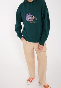 Grøn hoodie med et lilla kattegrafik og "Volcom"-bogstaver; beige plisserede bukser; orange sneakers; afslappet streetwear stil.