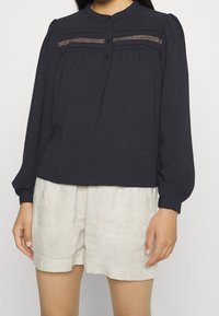 Blouse bleu marine foncé à manches longues avec col rond, boutonnage sur le devant et accents de couture horizontale texturée. Portée avec un short beige clair.