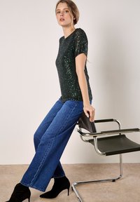 A model leans against a modern chair, wearing a sparkly green top and wide-leg jeans, with black heels and soft, wavy hair.