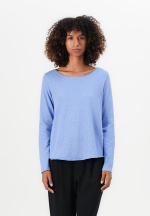 Femme avec des cheveux bouclés portant un haut à manches longues bleu clair et un pantalon noir, se tenant contre un fond blanc uni.